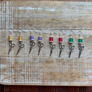 May include: A set of eight earrings featuring a miniature spool of thread and a pair of scissors. The spools are in various colors, including yellow, purple, red, and green.