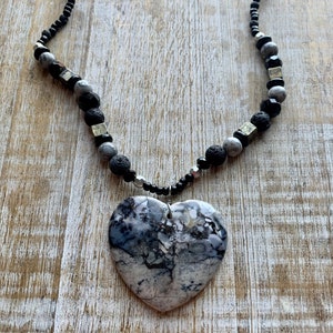 May include: A heart-shaped pendant necklace. The pendant is a large, marbled stone in shades of gray and white. The necklace features a black beaded chain with accents of gray, black, and metallic beads. The necklace is displayed on a wooden surface.
