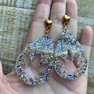 Op de afbeelding: Een paar oorhangers met een gouden hartbedel aan de bovenkant. De oorhangers zijn gemaakt van helder hars met gouden en blauwe glitter die in het hars zijn ingebed. Het hars is traanvormig met een bladvormige uitsnijding in het midden.