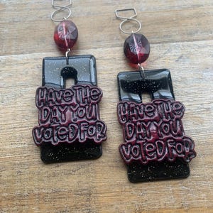 May include: A pair of black glitter earrings with the text "Have the day you voted for" in burgundy. Each earring has a rectangular shape with a cutout and a red bead at the top, connected by a silver hook.