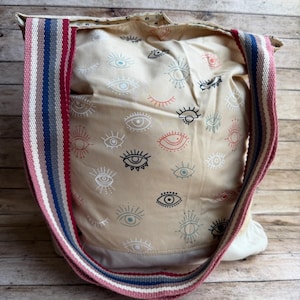 May include: Beige tote bag with an all-over eye print in white, red, and black. The bag has a striped shoulder strap in pink, blue, and gray. The bag is made of fabric and appears to be a shoulder bag.