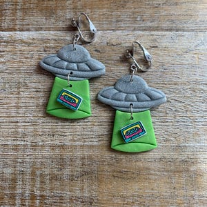 May include: A pair of silver clip-on earrings featuring a green and blue cassette tape charm dangling from a gray UFO charm.