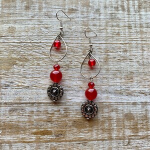 Op de afbeelding: Een paar oorhangers met rode glazen kralen en zilveren metalen accenten. De oorhangers hebben een traanvormig ontwerp met een kleine kraal aan de bovenkant en een grotere kraal aan de onderkant. De kralen zijn verbonden door een zilveren metalen ketting.