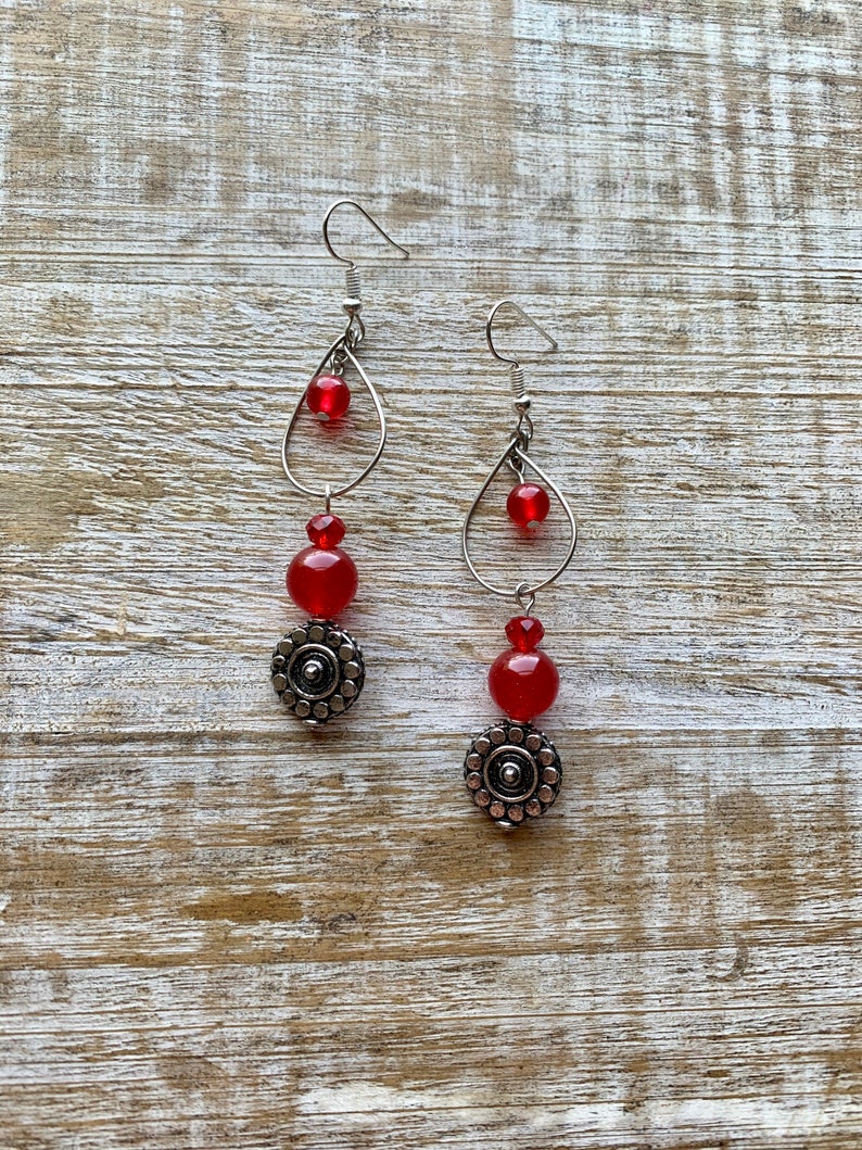 Op de afbeelding: Een paar oorhangers met rode glazen kralen en zilveren metalen accenten. De oorhangers hebben een traanvormig ontwerp met een kleine kraal aan de bovenkant en een grotere kraal aan de onderkant. De kralen zijn verbonden door een zilveren metalen ketting.