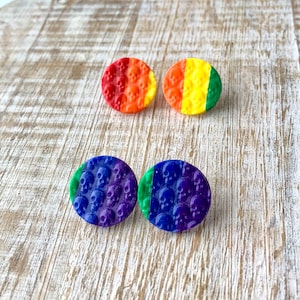 Puede incluir: Un conjunto de cuatro pendientes de calavera arcoíris. Los pendientes están hechos de arcilla polimérica y tienen forma de calavera. Las calaveras están pintadas en colores arcoíris, con cada color representando una parte diferente del arcoíris. Los pendientes son una forma divertida y única de mostrar tu orgullo.