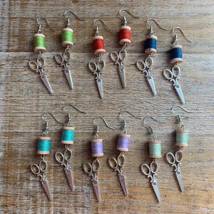 May include: A set of 10 pairs of earrings featuring a miniature spool of thread and a pair of scissors. The spools of thread are in various colors, including green, red, blue, turquoise, purple, and light green. The scissors are silver.