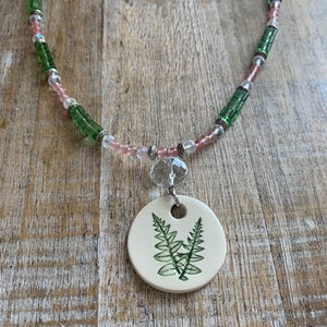 May include: A green and pink beaded necklace with a ceramic pendant featuring a green fern design.