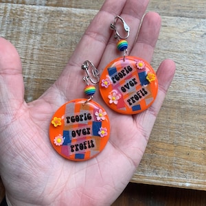 May include: Orange circular earrings with the text "people over profit". The earrings have a collage design with colorful squares and small flower accents. Each earring has a clip-on closure and a rainbow bead.
