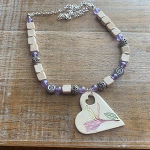 Puede incluir: Collar hecho a mano con un colgante en forma de corazón con un diseño floral. El collar tiene una cadena plateada y está adornado con cuentas cuadradas de madera, cuentas moradas y cuentas plateadas decorativas. El colgante es blanco con una flor rosa y verde pintada a mano.