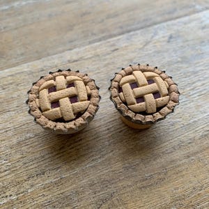 May include: Two miniature pie-shaped earrings. Each earring features a lattice crust design with a dark red filling, set in a metal cup. The earrings are placed on a wooden surface.