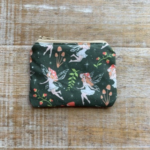 May include: A small green zippered pouch with a whimsical fairy and mushroom print. The pouch features a repeating pattern of fairies with wings, mushrooms, and stars on a dark green background.
