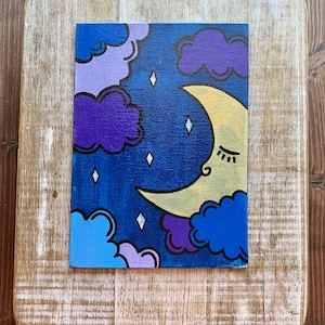 May include: A hand-painted canvas with a crescent moon and clouds in shades of purple, blue, and yellow against a dark blue background. The moon has a closed eye and a small smile.