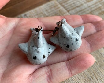 May include: A pair of gray and white shark earrings with black spots. The earrings are made of polymer clay and have a dangling design.