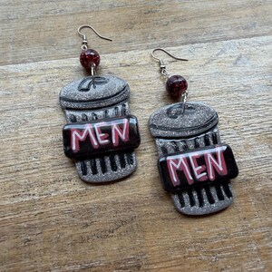 May include: A pair of novelty earrings featuring a coffee cup design. The earrings are gray with silver glitter accents and a black band with the word "MEN" in white and red. Each earring has a red bead and silver hook.