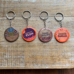 Puede incluir: Cuatro llaveros con botones redondos de color naranja y marrón. Cada botón tiene un mensaje diferente: "Boycott Bezos", "Boycott Elon", "Boycott Corporations" y "Boycott Facebook".