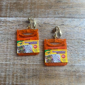 May include: Orange Crayola colored pencil box earrings with gold-colored clip-on findings. Each earring features a miniature box with the text "Create & Color" and "65" on the front. The earrings are set against a wooden background.