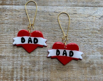 Könnte beinhalten: Ein Paar rote herzförmige Ohrringe mit weißen Bannern, auf denen in schwarzen Buchstaben "DAD" steht. Die Ohrringe sind an goldenen Kreisen befestigt.