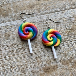 Puede incluir: Un par de pendientes de piruleta coloridos. Cada pendiente presenta un diseño de remolino arcoíris en tonos de rojo, naranja, amarillo, verde, azul y morado. Las piruletas están unidas a ganchos para la oreja de color plateado con palitos blancos.