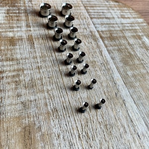 May include: A set of 14 silver metal ear gauges of varying sizes. The gauges are arranged in a diagonal line on a light brown wooden surface.