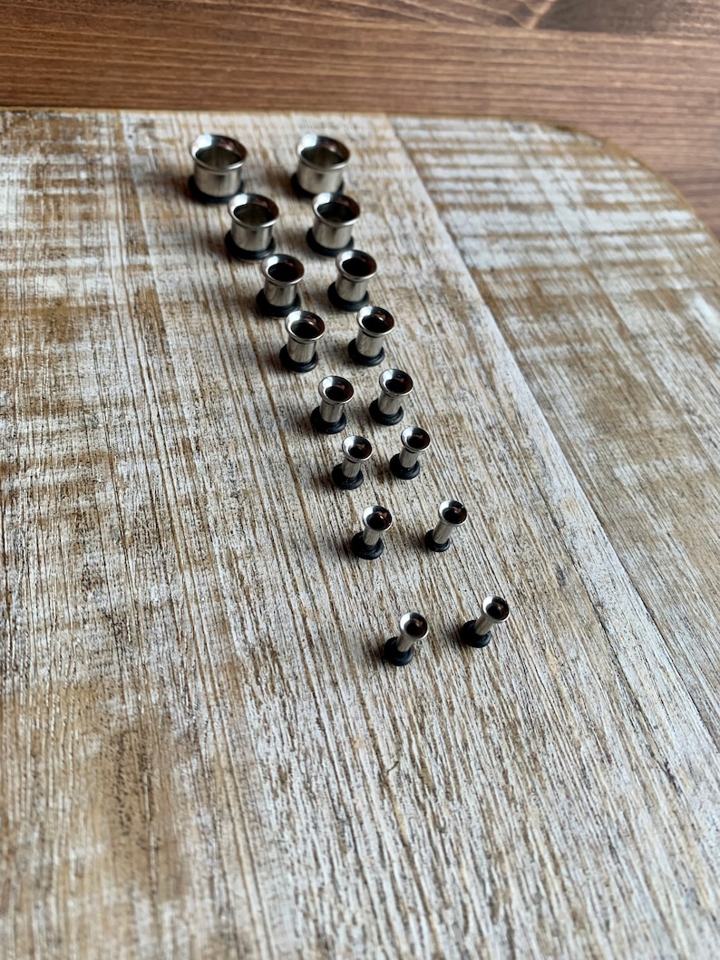 May include: A set of 14 silver metal ear gauges of varying sizes. The gauges are arranged in a diagonal line on a light brown wooden surface.