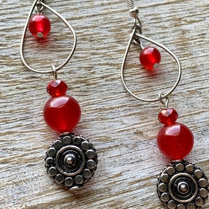 Op de afbeelding: Rode en zilveren oorhangers met een traanvorm en een ronde kraal. De oorhangers hebben een zilveren bedel met een cirkelvormig ontwerp.