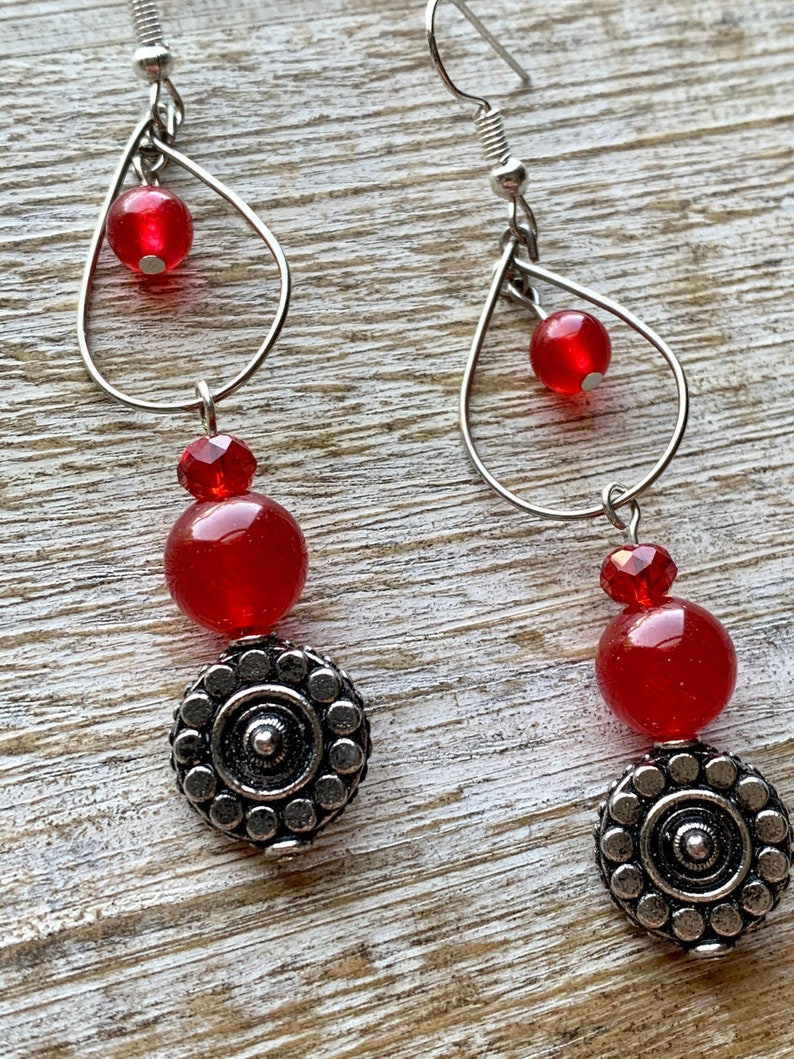 Op de afbeelding: Rode en zilveren oorhangers met een traanvorm en een ronde kraal. De oorhangers hebben een zilveren bedel met een cirkelvormig ontwerp.