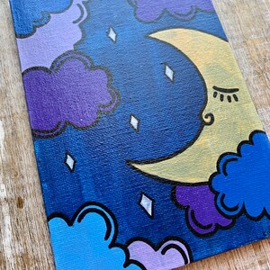 May include: A hand-painted canvas featuring a crescent moon with a closed eye and eyelashes, surrounded by purple, blue, and light blue clouds on a dark blue background. The moon is yellow with a white outline. The clouds have black outlines. There are white diamond-shaped stars scattered across the canvas.