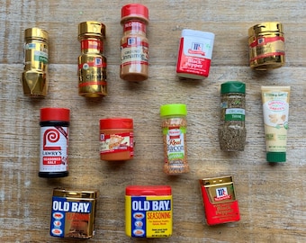 May include: A selection of miniature spice jars and seasoning containers, including Old Bay and McCormick spices, arranged on a wooden surface. The jars display a variety of colours and labels, with some containing ground cinnamon and black pepper.