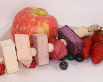 May include: A wooden skewer with colourful fruit and wood pieces. The skewer has a rope attached to one end. The skewer is on a white plate with a red apple, strawberries, and blueberries.