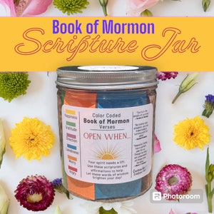 May include: A glass jar with a silver lid filled with colorful paper strips. The jar is labeled "Color Coded Book of Mormon Verses" and "OPEN WHEN..." The paper strips have words like "Happiness", "Gratitude", "Direction", "Loneliness", "Worry", and "Angry" printed on them. The jar is surrounded by flowers.