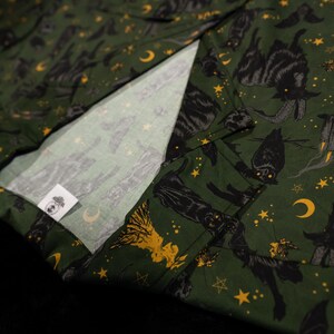 Puede incluir: Tela verde oscuro con un patr&oacute;n repetido de gatos negros, lunas, estrellas y otros s&iacute;mbolos m&iacute;sticos. La tela tiene un interior blanco y una peque&ntilde;a etiqueta con un logotipo.