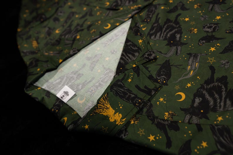 Puede incluir: Tela verde oscuro con un patr&oacute;n repetido de gatos negros, lunas, estrellas y otros s&iacute;mbolos m&iacute;sticos. La tela tiene un interior blanco y una peque&ntilde;a etiqueta con un logotipo.