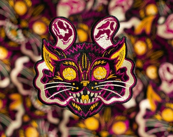 May include: A black and yellow embroidered patch of a cat with a spooky, grinning face. The cat has large, round eyes with yellow pupils and a wide, toothy grin. The cat's ears are shaped like ghosts with white outlines and a purple interior. The patch is surrounded by a white outline and has a white, wispy cloud effect around the cat's head.