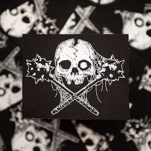 May include: Black square patch featuring a white skull with a cracked forehead, crossed maces, and spiderwebs. The patch has a gothic or punk rock aesthetic. The background is a black fabric with a repeating skull pattern.