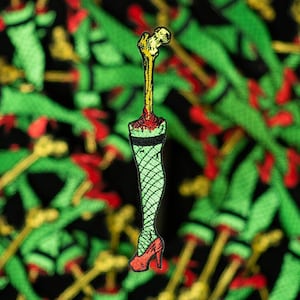 May include: A black and green embroidered patch of a leg in a fishnet stocking with a bone sticking out of the top, wearing a red high heel shoe.