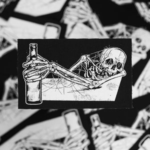 May include: A black and white embroidered patch of a skeleton holding a bottle of alcohol. The skeleton is lying down and has a web-like design around it.