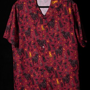 Puede incluir: Camisa abotonada burdeos con un estampado repetido de cabras negras, gatos, murci&eacute;lagos, lunas, estrellas y figuras. La camisa de manga corta tiene un cuello cl&aacute;sico y un bolsillo en el pecho.