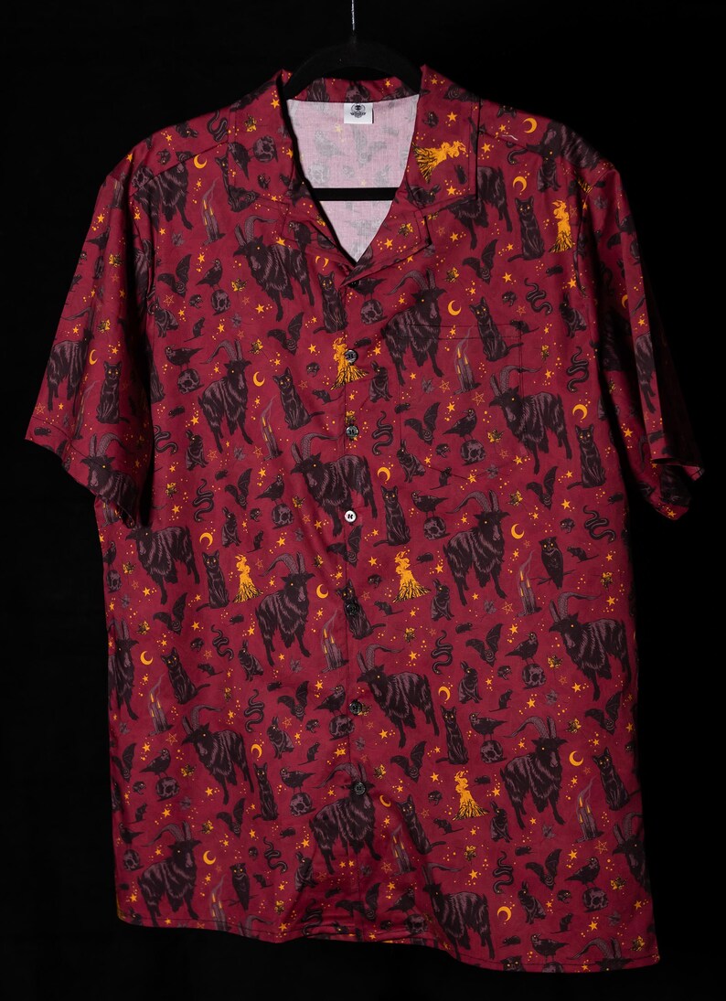 Puede incluir: Camisa abotonada burdeos con un estampado repetido de cabras negras, gatos, murci&eacute;lagos, lunas, estrellas y figuras. La camisa de manga corta tiene un cuello cl&aacute;sico y un bolsillo en el pecho.