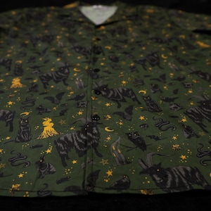 Puede incluir: Camisa verde oscuro con botones y un estampado repetido de gatos negros, murci&eacute;lagos, serpientes, b&uacute;hos, estrellas y otros s&iacute;mbolos ocultos en amarillo. La camisa tiene cuello de campamento y botones negros.