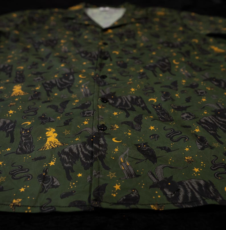 Puede incluir: Camisa verde oscuro con botones y un estampado repetido de gatos negros, murci&eacute;lagos, serpientes, b&uacute;hos, estrellas y otros s&iacute;mbolos ocultos en amarillo. La camisa tiene cuello de campamento y botones negros.