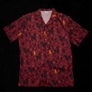 Puede incluir: Camisa de manga corta de color rojo oscuro, estampada con cabras, gatos, murci&eacute;lagos, lunas y estrellas negras. La camisa tiene un cuello cl&aacute;sico y un corte relajado, perfecta para el uso diario.