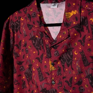 Puede incluir: Camisa burdeos con botones y un estampado negro y dorado de gatos, murci&eacute;lagos, lunas, estrellas y otros s&iacute;mbolos ocultos. La camisa tiene cuello de campamento y un bolsillo en el pecho. La camisa est&aacute; colgada en una percha negra.
