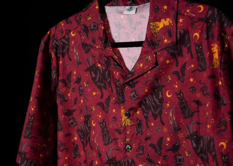 Puede incluir: Camisa burdeos con botones y un estampado negro y dorado de gatos, murci&eacute;lagos, lunas, estrellas y otros s&iacute;mbolos ocultos. La camisa tiene cuello de campamento y un bolsillo en el pecho. La camisa est&aacute; colgada en una percha negra.