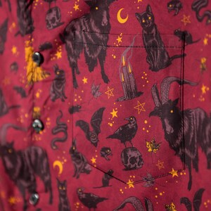 Puede incluir: Camisa abotonada burdeos con un estampado repetido de gatos negros, cabras, murci&eacute;lagos, serpientes, lunas, estrellas y velas. La camisa tiene un bolsillo y botones negros.