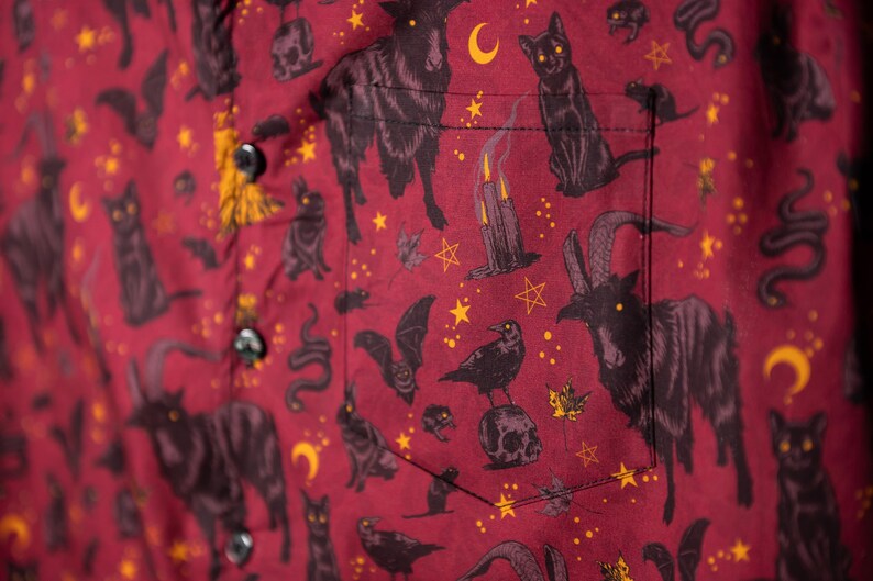 Puede incluir: Camisa abotonada burdeos con un estampado repetido de gatos negros, cabras, murci&eacute;lagos, serpientes, lunas, estrellas y velas. La camisa tiene un bolsillo y botones negros.