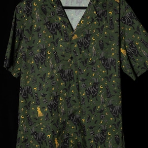 Puede incluir: Camisa verde con botones con un estampado repetido de cabras negras, gatos, murci&eacute;lagos, lunas, estrellas y velas. La camisa de manga corta tiene cuello de campamento y est&aacute; hecha de tela ligera.