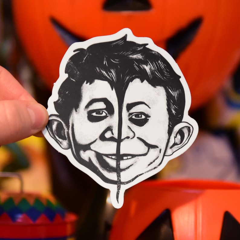 May include: Black and white sticker of a cartoon character with a mischievous grin. The character has a black line down the center of its face, dividing the left and right sides.