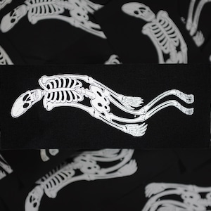 May include: A black rectangular patch featuring a white, stylized skeleton design. The skeleton appears to be in motion, with elongated limbs and a curved spine. The patch is likely for clothing or accessories.