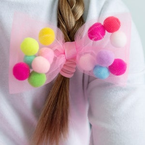 May include: A pink tulle bow with colorful pom-poms, used as a hair tie.
