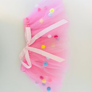 May include: A pink tulle tutu skirt with a white ribbon and colorful pom-poms.